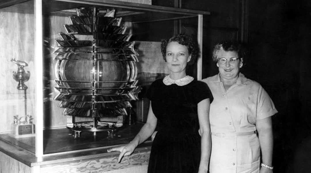 Two women standing in front of the Fresnel lens, which is inside a glass case on a pedestal.