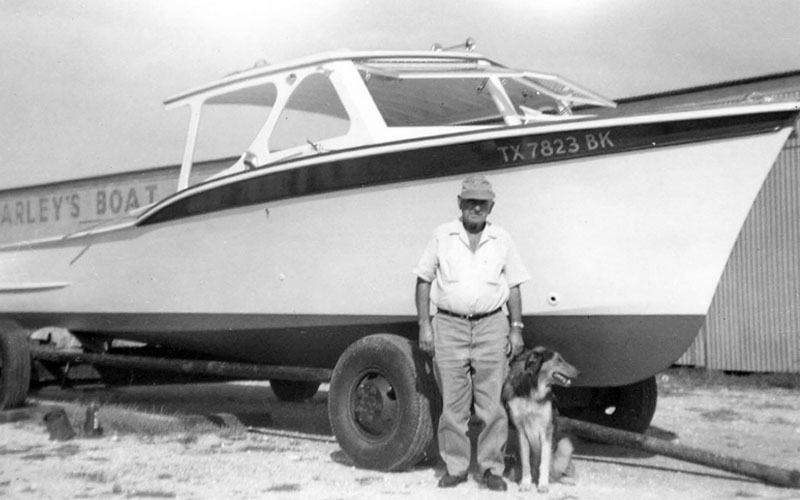 A man in a white shirt and a dog standing in front of a boat on a trailer.